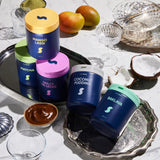Assorted dessert containers on a decorative tray with fruits and a coconut.