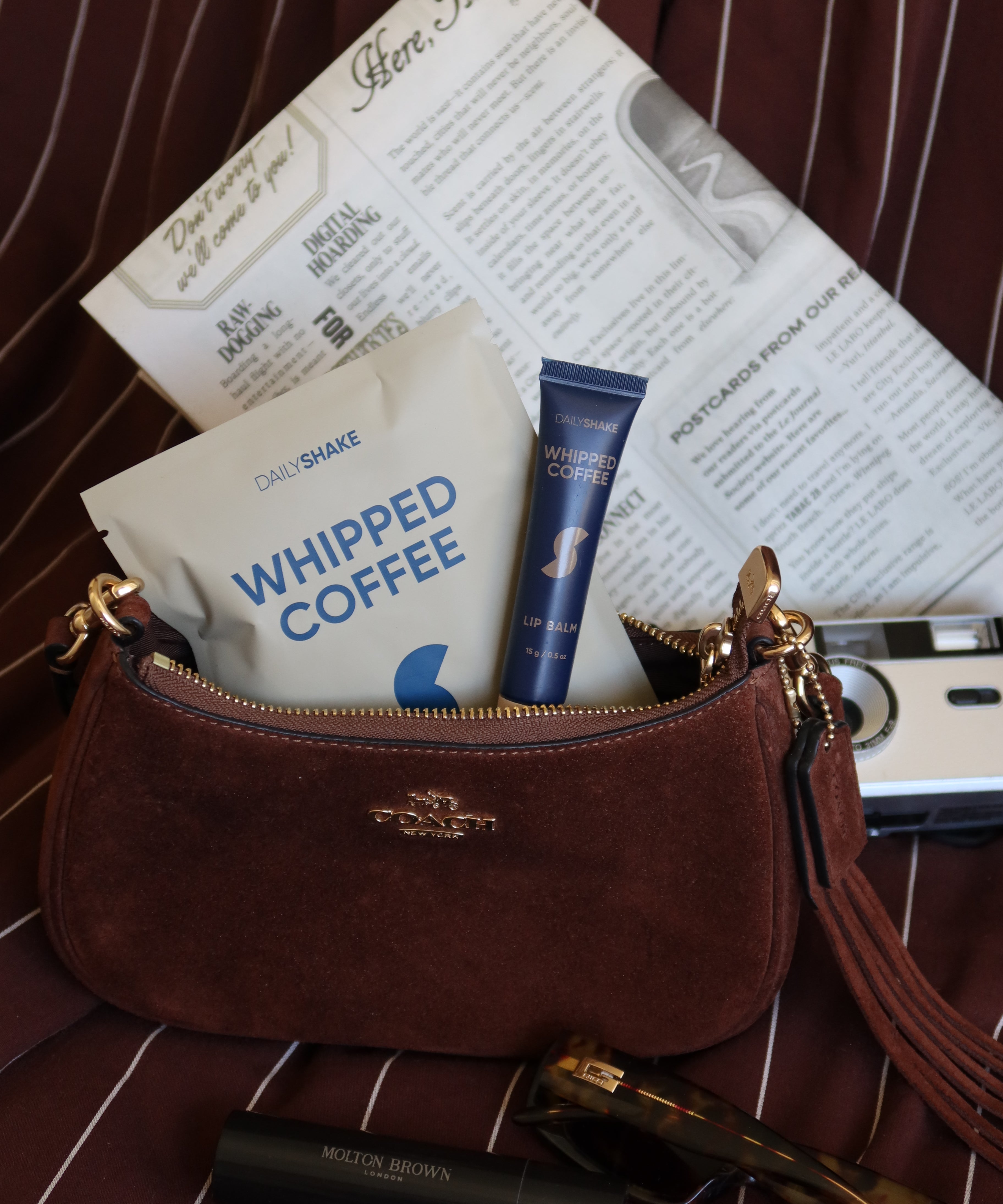 Brown handbag with cosmetic items including a tube of whipped coffee creamer on a striped surface.
